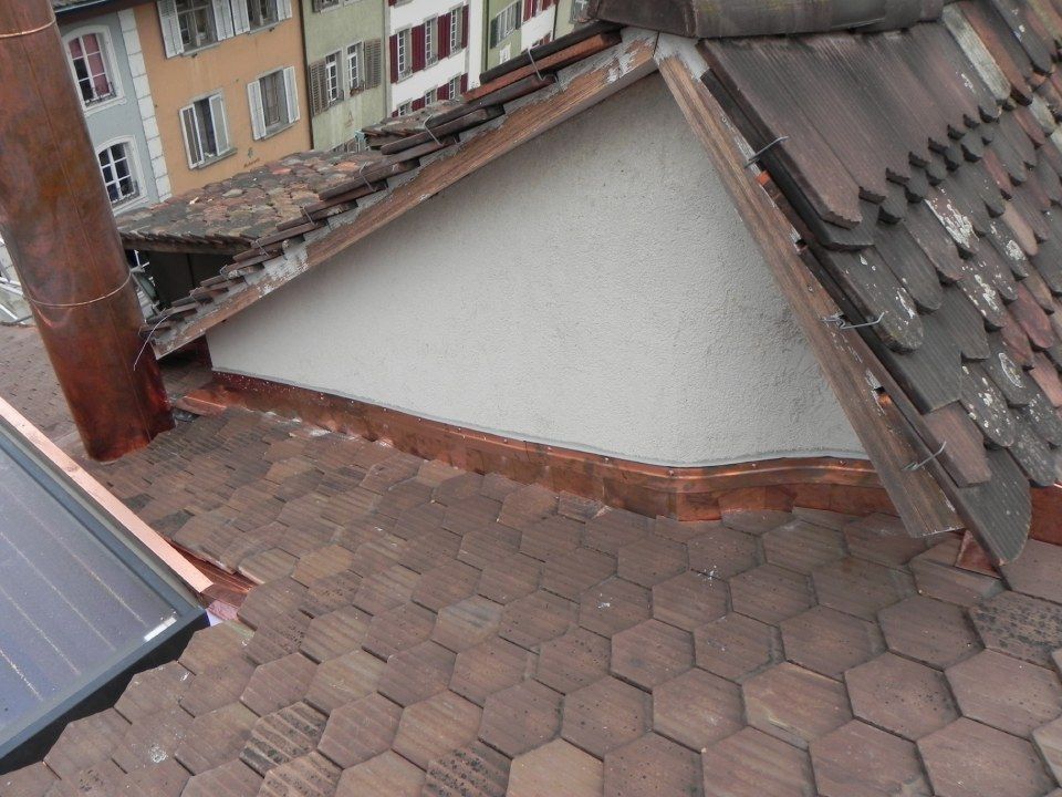 Nahaufnahme eines mit Schindeln gedeckten Daches mit einer verputzten Dachgaube, einem metallenen Schornsteinrohr und einem Solarpanel in der Ecke.