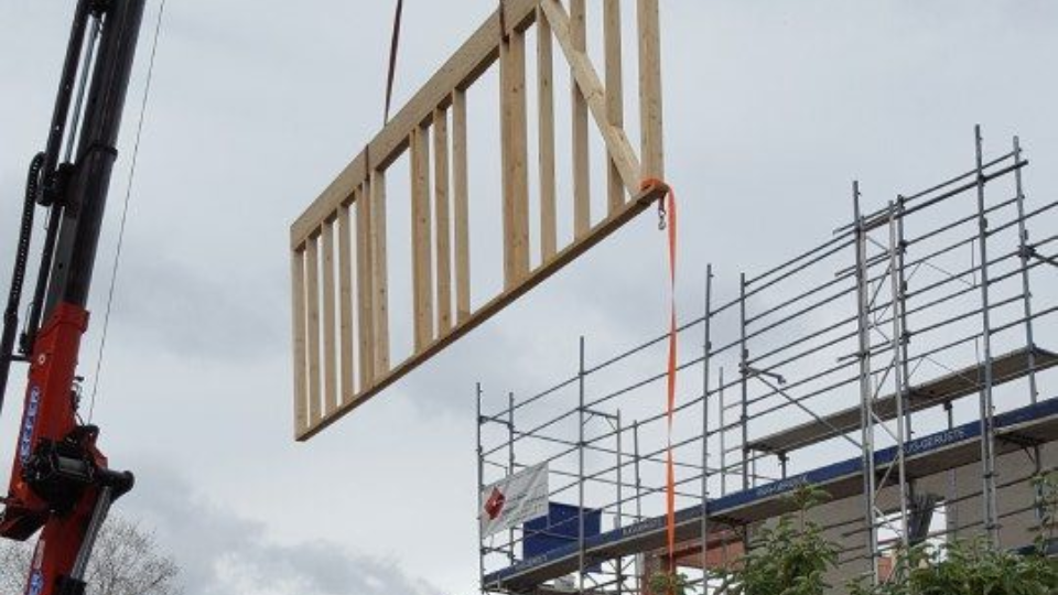 Auf einer Baustelle hebt ein LKW-Kran ein grosses hölzernes Wandgerüst an; im Hintergrund ist ein Baugerüst zu sehen.