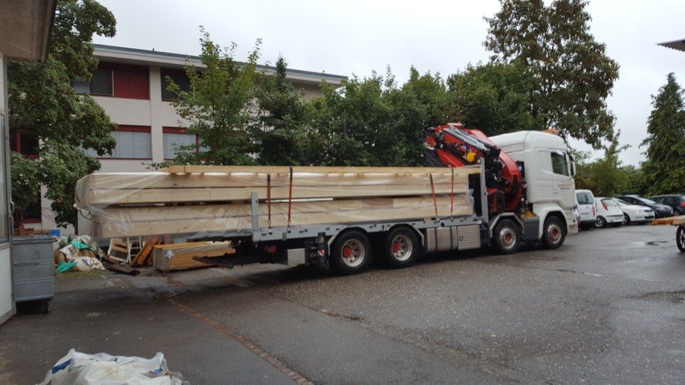 Ein mit langen Holzbalken beladener Pritschenwagen parkte auf einem gepflasterten Parkplatz neben einem Gebäude.