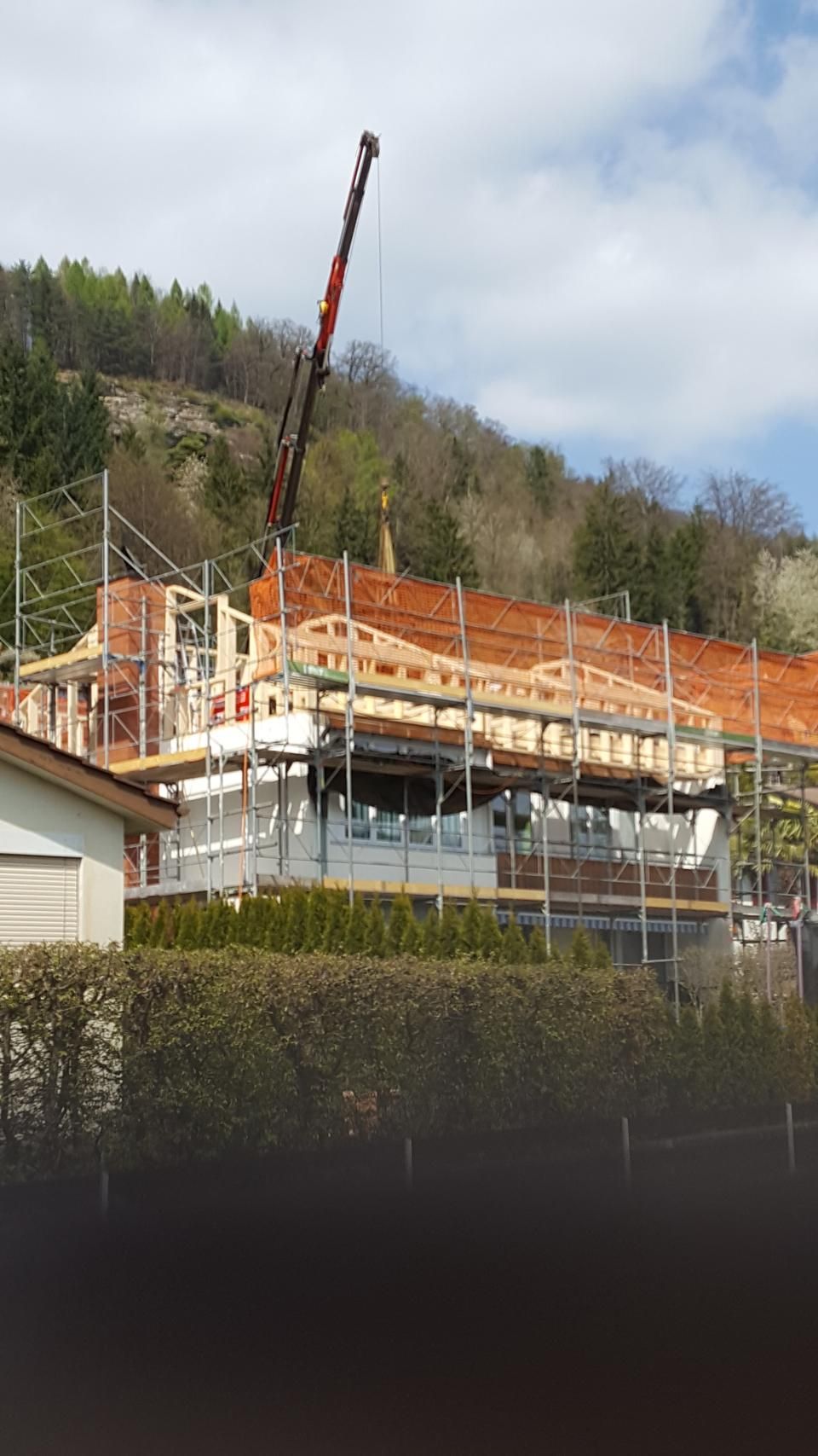 Ein im Bau befindliches Haus mit Gerüst, einem Kran und einem bewaldeten Hügel im Hintergrund unter blauem Himmel.