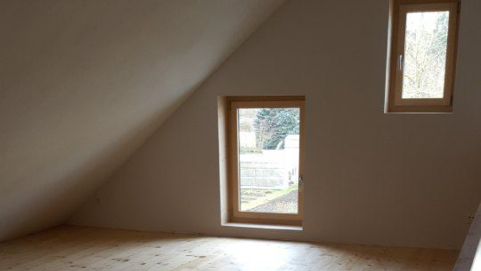 Innenansicht einer schrägen Decke mit einer Wand aus astigen Holzpaneelen und zwei kleinen rechteckigen Fenstern an einer schlichten Wand.