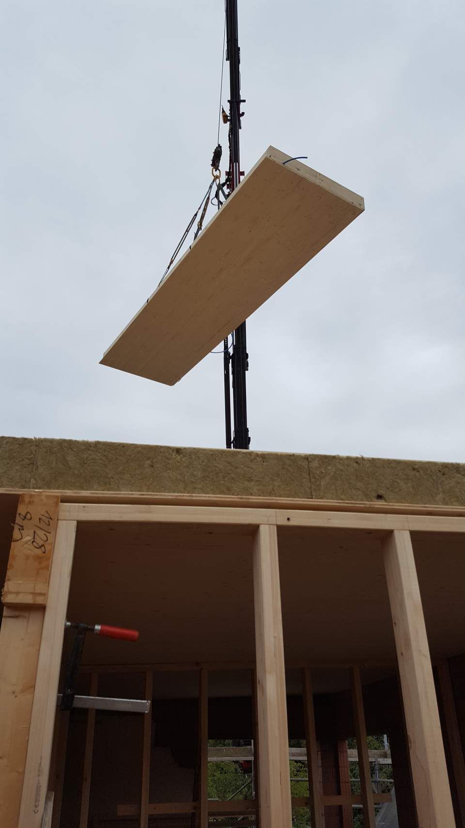 Ein Kran hebt eine grosse, rechteckige Holzplatte über die unfertige Holzrahmenkonstruktion eines Gebäudes.