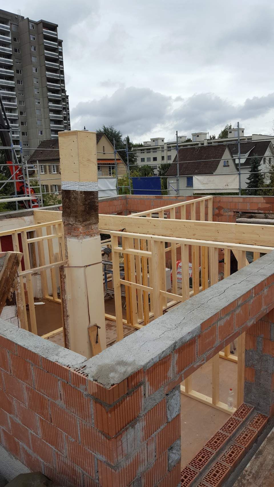 Auf einer Baustelle befindet sich eine Holzrahmenkonstruktion innerhalb von Ziegelmauern; in der Ferne sind Wohngebäude zu sehen.