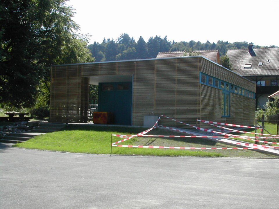 Ein modernes, holzverkleidetes Gebäude mit blauem Eingang, gelegen auf einer Rasenfläche in der Nähe von Bäumen, teilweise abgesperrt durch Absperrband.