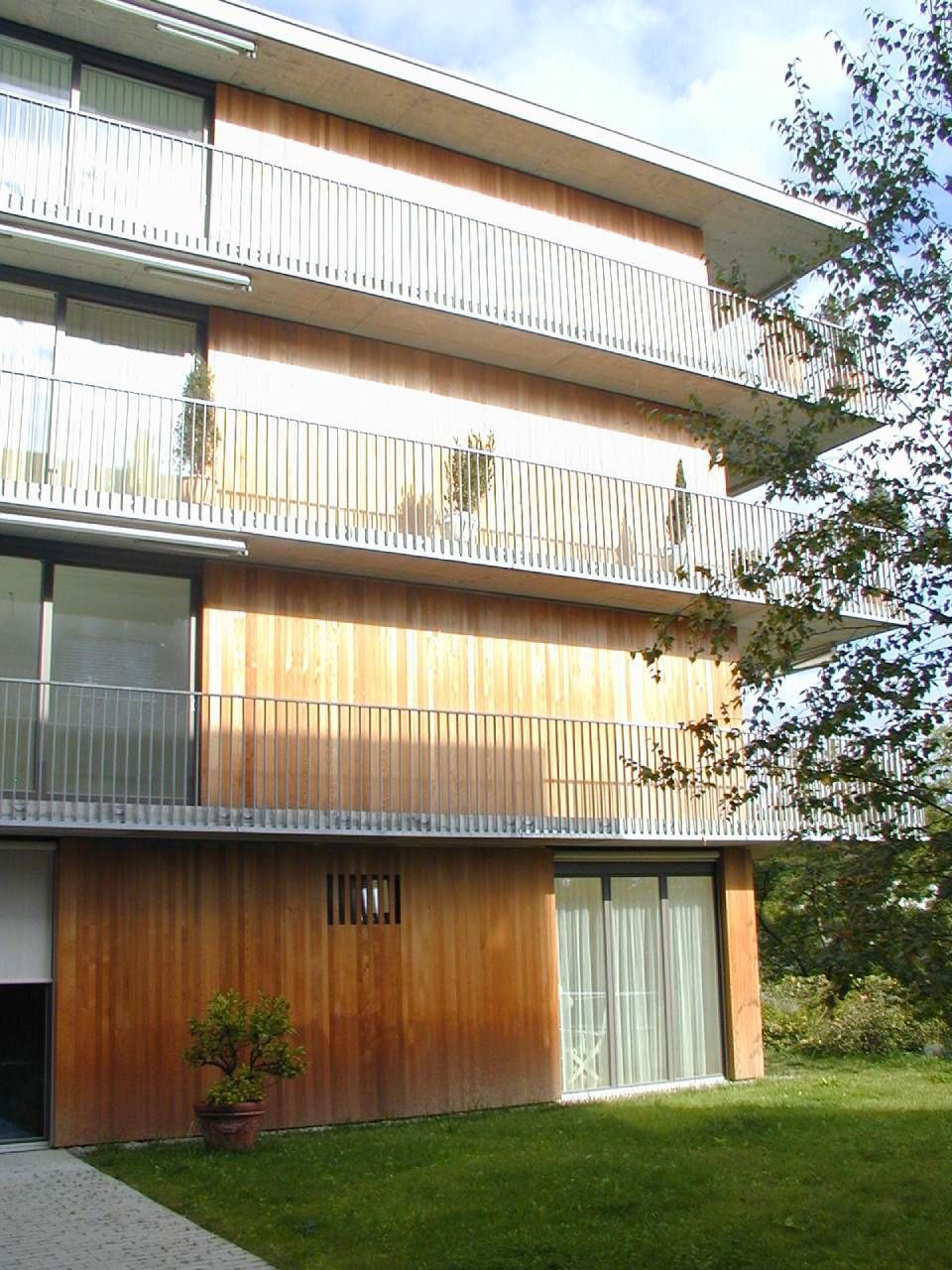 Ein modernes Apartmentgebäude mit holzgetäfelten Wänden, mehreren Etagen mit Balkonen mit Geländern und einer grünen Rasenfläche.