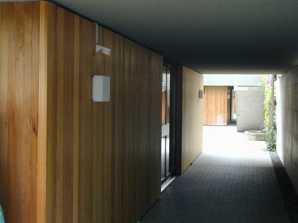 Links befindet sich ein moderner, überdachter Gehweg mit vertikaler Holzvertäfelung, der zu einem offenen, sonnigen Außenbereich führt.