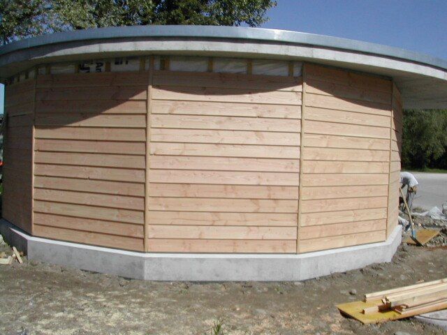 Ein im Bau befindliches kreisrundes Gebäude mit heller Holzplankenverkleidung auf einem grauen Betonfundament.