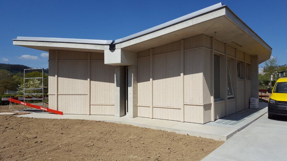 Ein modernes, hellfarbiges Modulgebäude mit markantem Schmetterlingsdach, gelegen auf einem Kiesplatz unter blauem Himmel.