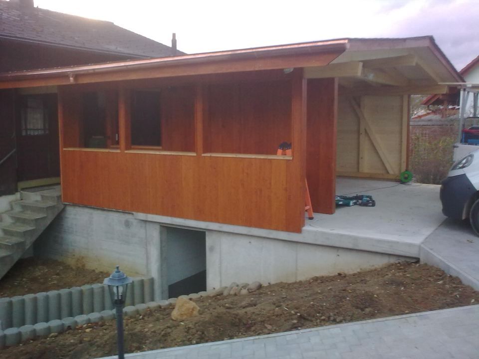 Eine teilweise fertiggestellte hölzerne Verandaerweiterung, die an ein Haus angebaut ist und über Betonfundamentwände und einen Carportbereich verfügt.
