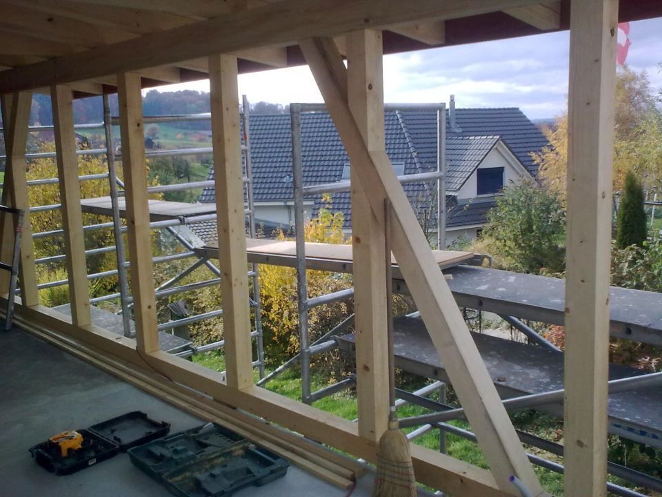 Eine im Bau befindliche Holzrahmenkonstruktion, von innen betrachtet, mit Blick auf eine Wohngegend mit Baugerüsten.