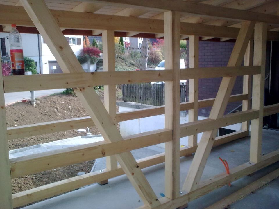 Auf einem Betonboden steht eine Holzrahmenkonstruktion mit diagonalen Verstrebungsbalken; im Hintergrund ist der Aussenbereich verschwommen dargestellt.