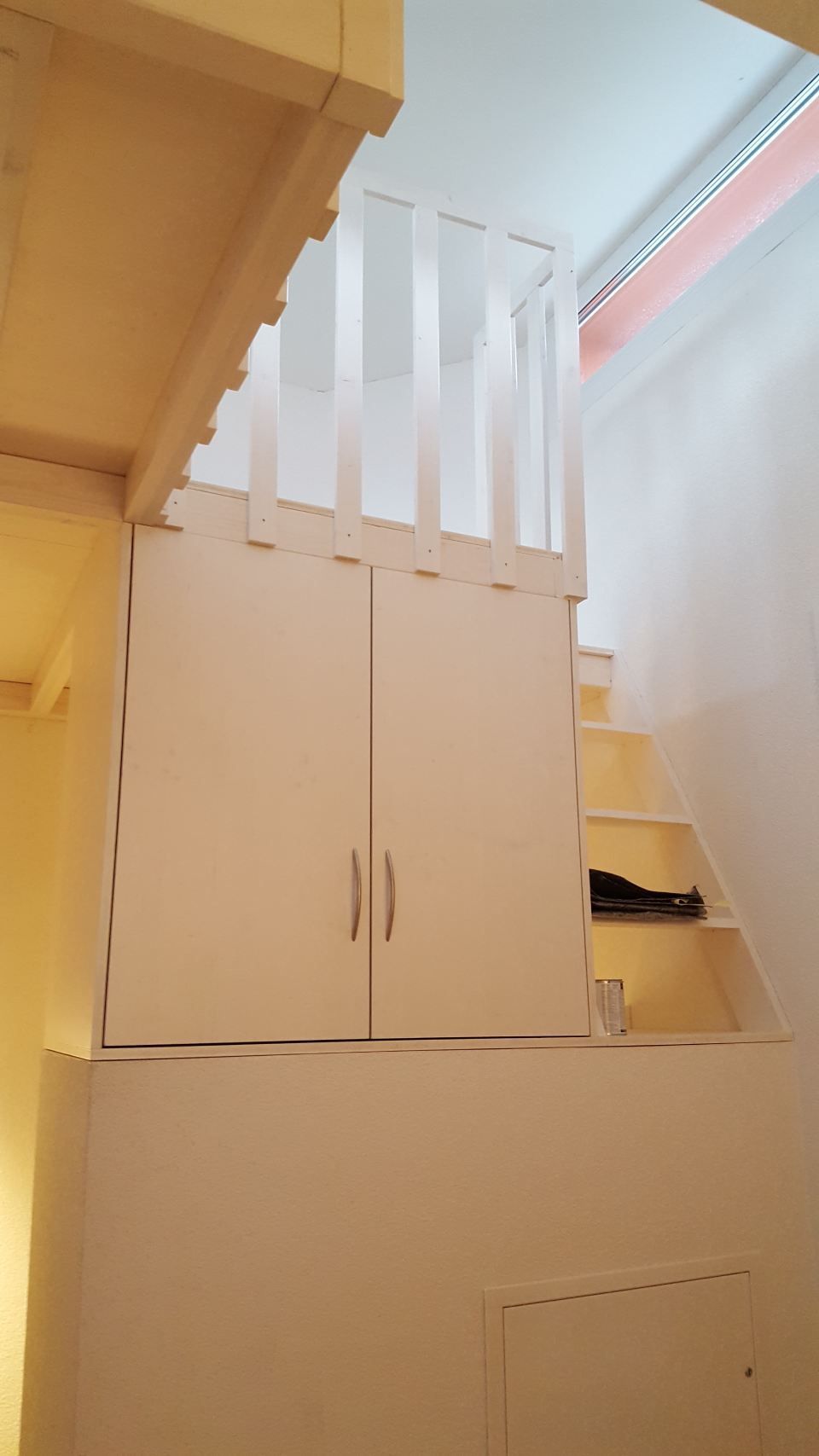 Maßgefertigter, weisser Aufbewahrungsschrank mit eingebauten Schränken und Treppe, die zu einem oberen Loftbereich mit einem schlichten weissen Geländer führt.
