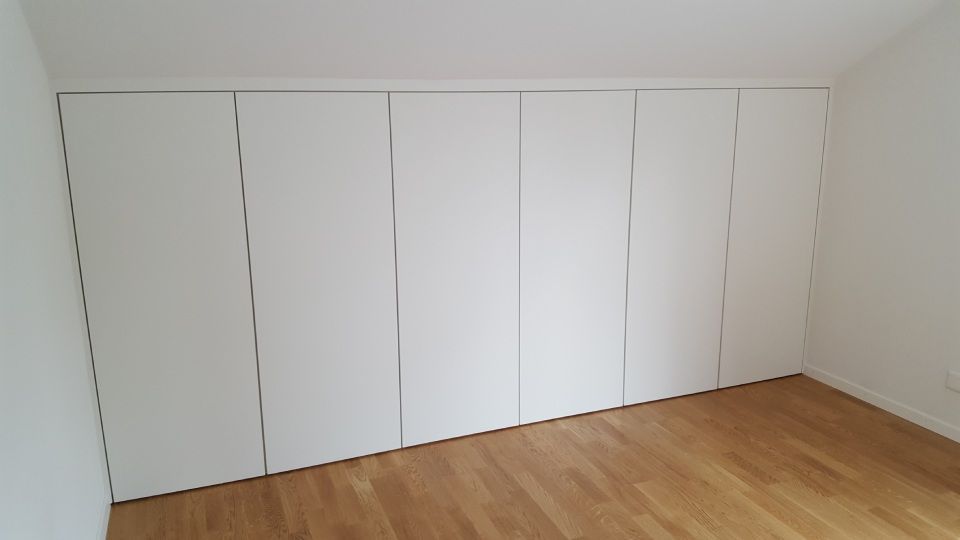Eine Wand aus weissen, raumhohen Einbauschränken mit minimalistischen vertikalen Griffen vor einem Holzboden.
