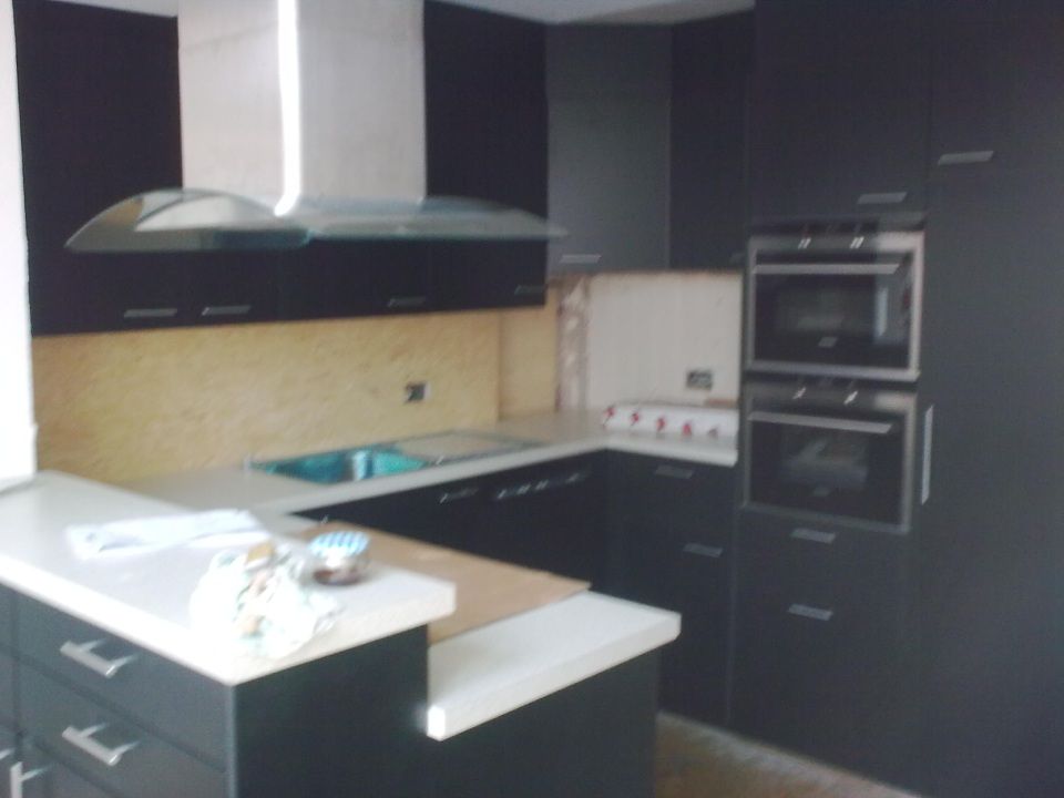 Eine moderne Küche mit schwarzen Schränken, weissen Arbeitsplatten, einer Dunstabzugshaube aus Edelstahl und übereinander angeordneten Wandbacköfen.