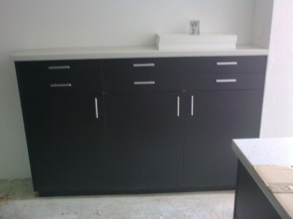 Ein dunkelbrauner Schrank mit Schubladen und Fächern, darauf eine weisse Arbeitsplatte und ein kleines Waschbecken vor einer weissen Wand.