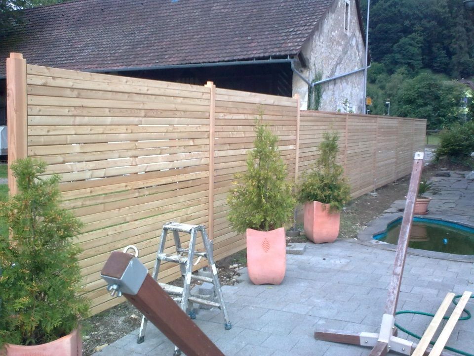 In einem Wohngarten steht ein neuer Sichtschutzzaun aus Holz auf einer Steinterrasse, daneben befinden sich Topfpflanzen aus immergrünem Laub und eine Leiter.