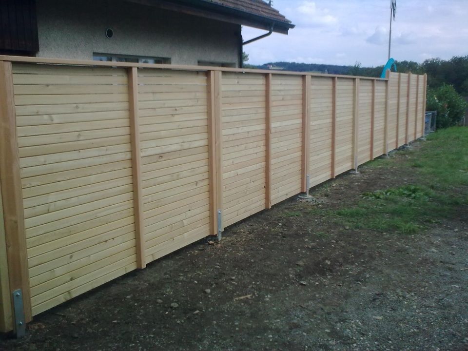 Ein langer, neuer Sichtschutzzaun aus Holz mit horizontalen Latten wurde entlang eines grasbewachsenen Hinterhofs auf Metallpfostenankern errichtet.