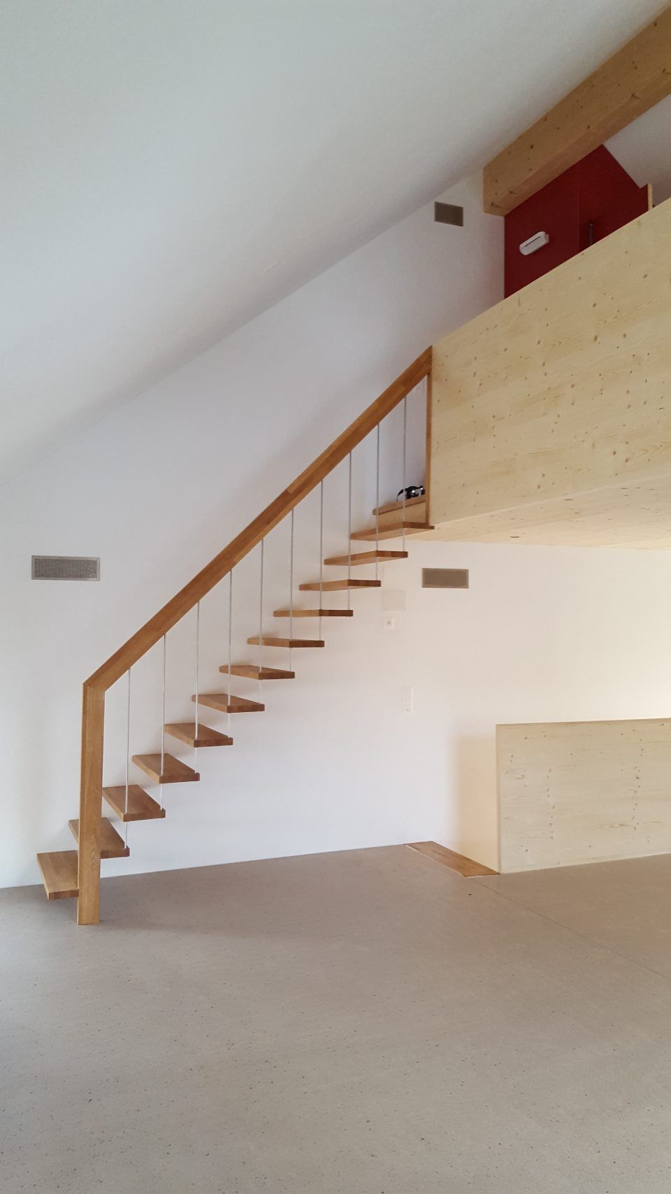 Minimalistisches Interieur mit einer freitragenden Holztreppe, die zu einem loftartigen Bereich vor einer weißen Wand führt.