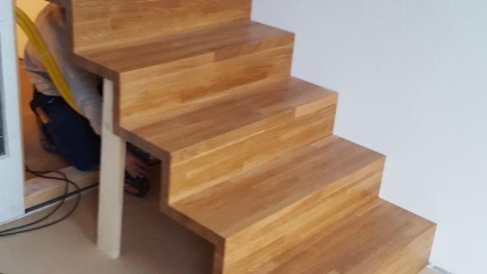 Eine Treppe mit hölzernen, offenen Stufen, die sich an einer schlichten weissen Wand nach oben winden.