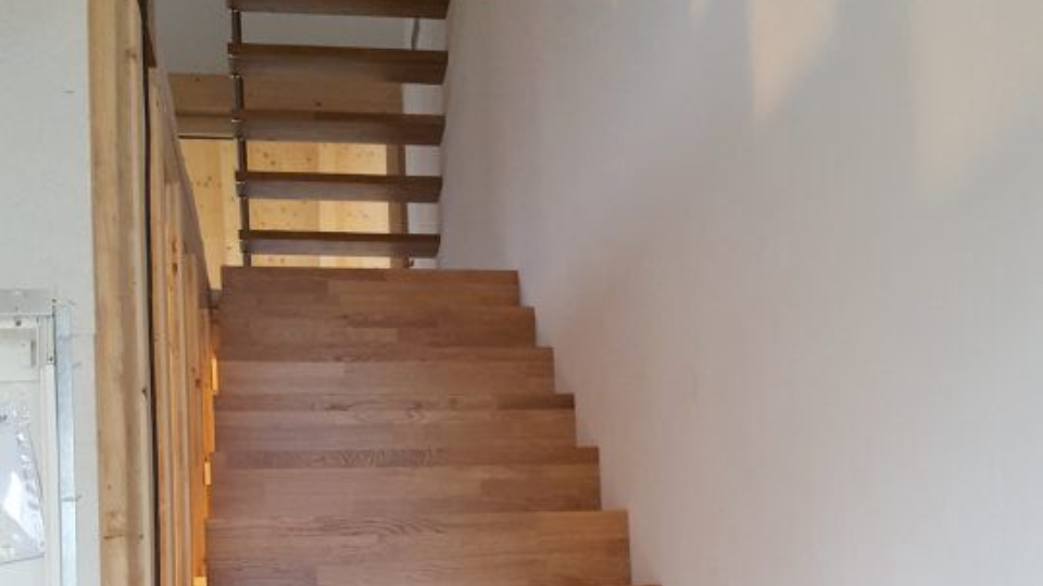 Eine helle Holztreppe mit vertikaler Paneelverkleidung führt hinauf zu einem Treppenabsatz mit Holzgeländer.