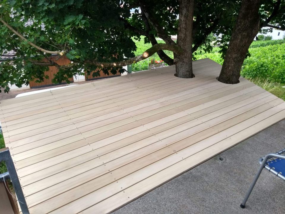 Eine um zwei Baumstämme herum gebaute Holzterrasse, teilweise von Ästen beschattet, mit Blick auf einen grünen Garten.