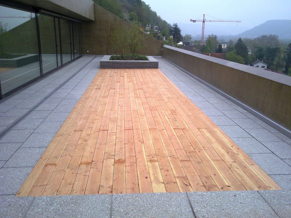 Eine rechteckige Holzterrasse innerhalb einer mit Steinplatten belegten Terrasse mit einem Hochbeet und Blick auf die Hügel im Hintergrund.