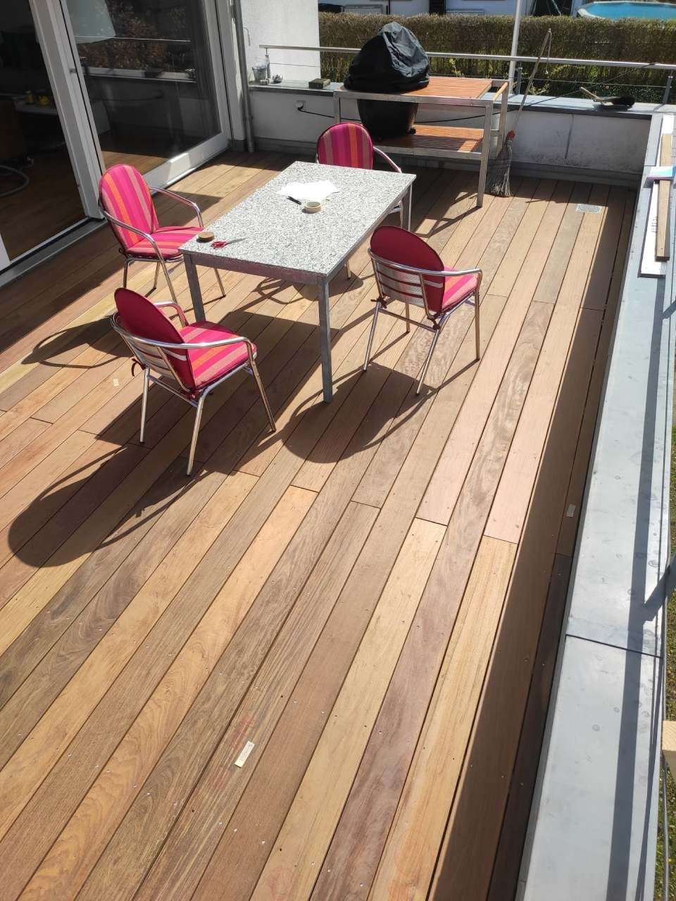 Eine Holzterrasse mit einem Steintisch und vier rosa gestreiften Stühlen, vor einer Terrassentür und einem Metallgeländer.