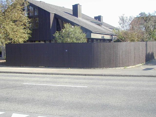 Entlang einer gepflasterten Strasse verläuft ein dunkelbrauner Holzzaun vor einem modernen Haus mit steilem Dach und dunkler Fassadenverkleidung.