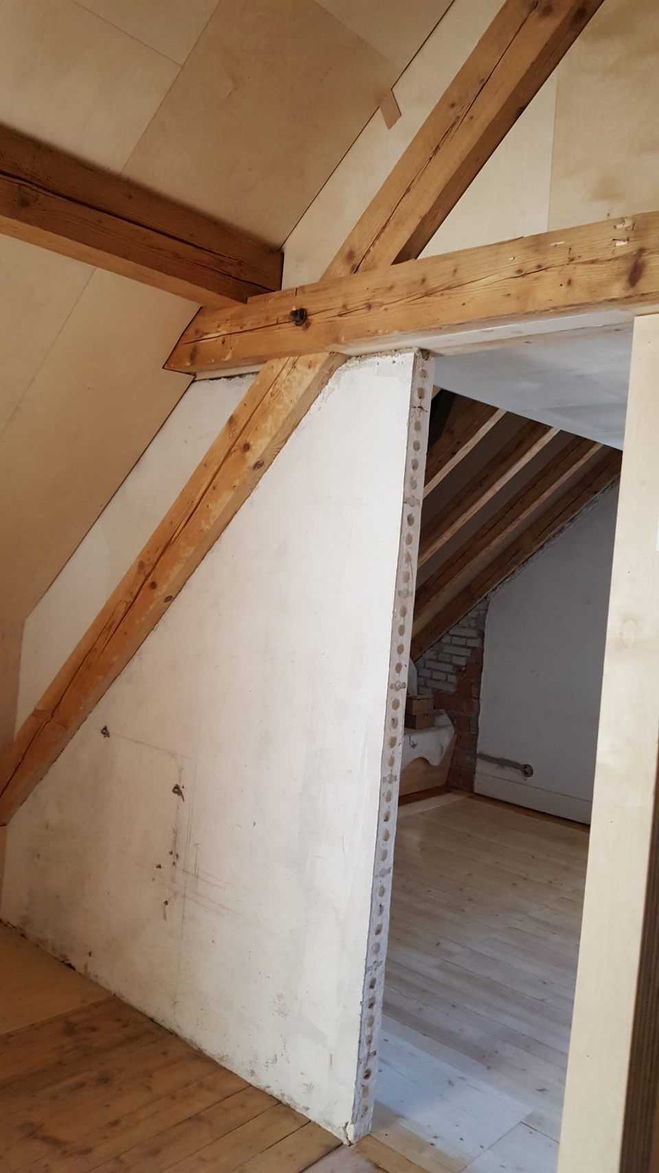 Ein Holzbalken verläuft schräg nach unten entlang einer weissen Putzwand, die zwei Räume in einem unausgebauten Dachgeschoss trennt.