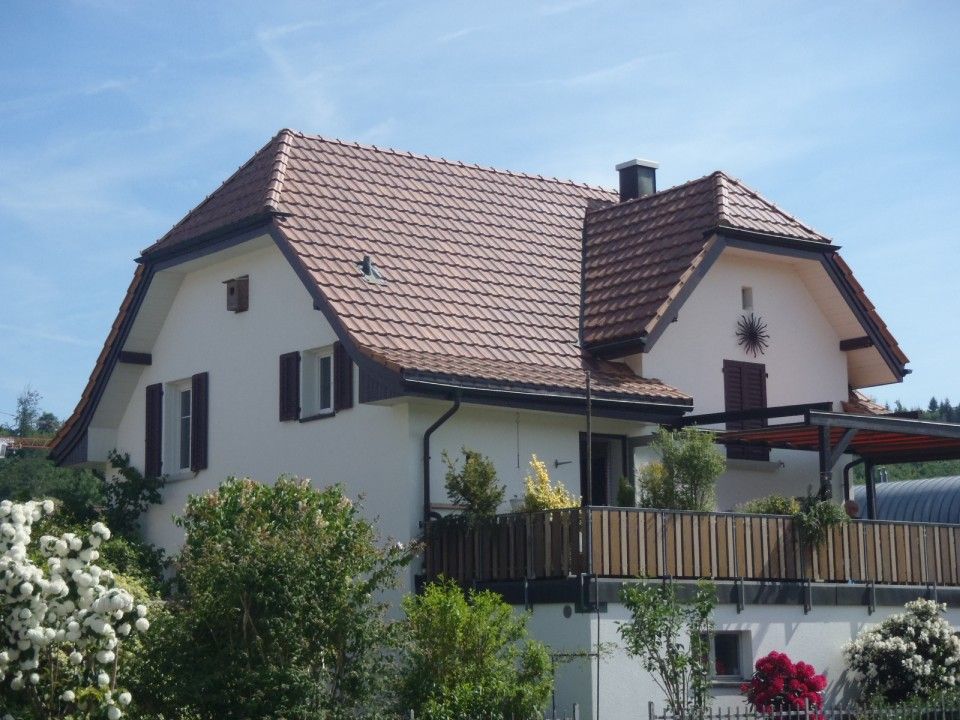 Ein zweistöckiges weisses Haus mit einem rot gedeckten, mehrfach geneigten Dach, dunkelbraunen Fensterläden und einem hölzernen Balkon an der Vorderseite.