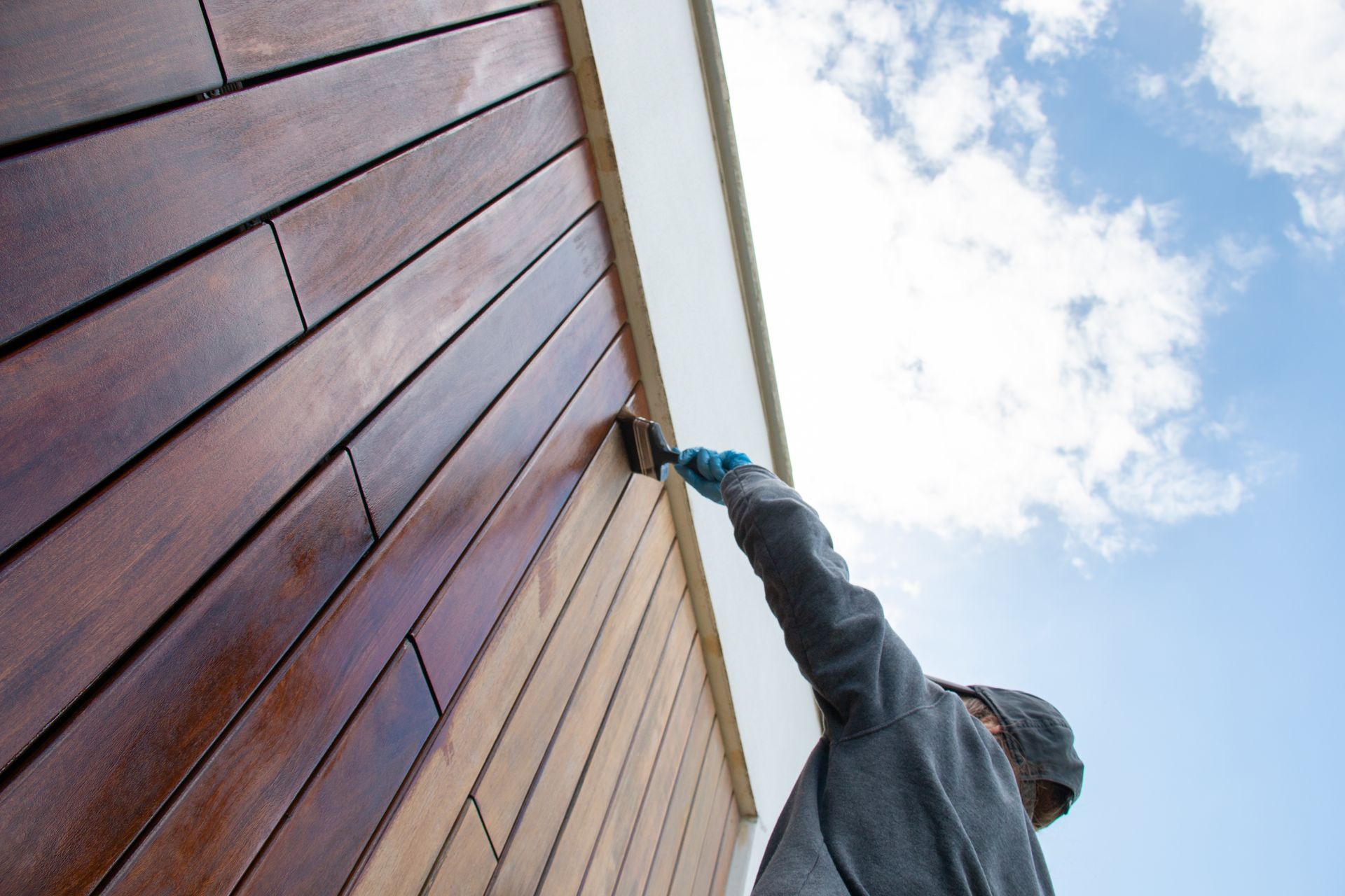 Une personne applique une teinture sur un mur en bois sous un ciel bleu.