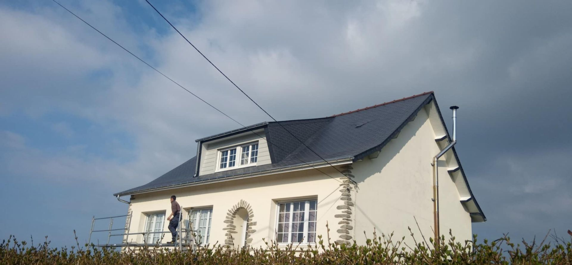 Une personne debout sur un échafaudage, travaillant sur une maison à deux étages au toit sombre, se détachant sur un ciel nuageux.