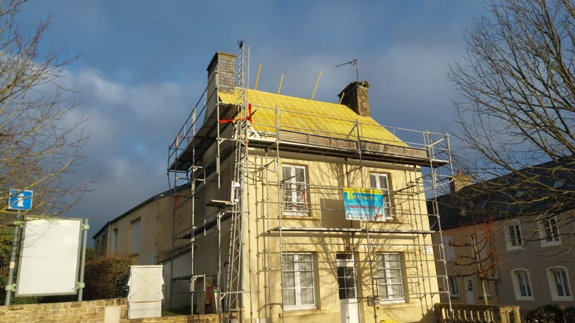 Maison en rénovation avec échafaudages, toiture jaune et ciel nuageux.