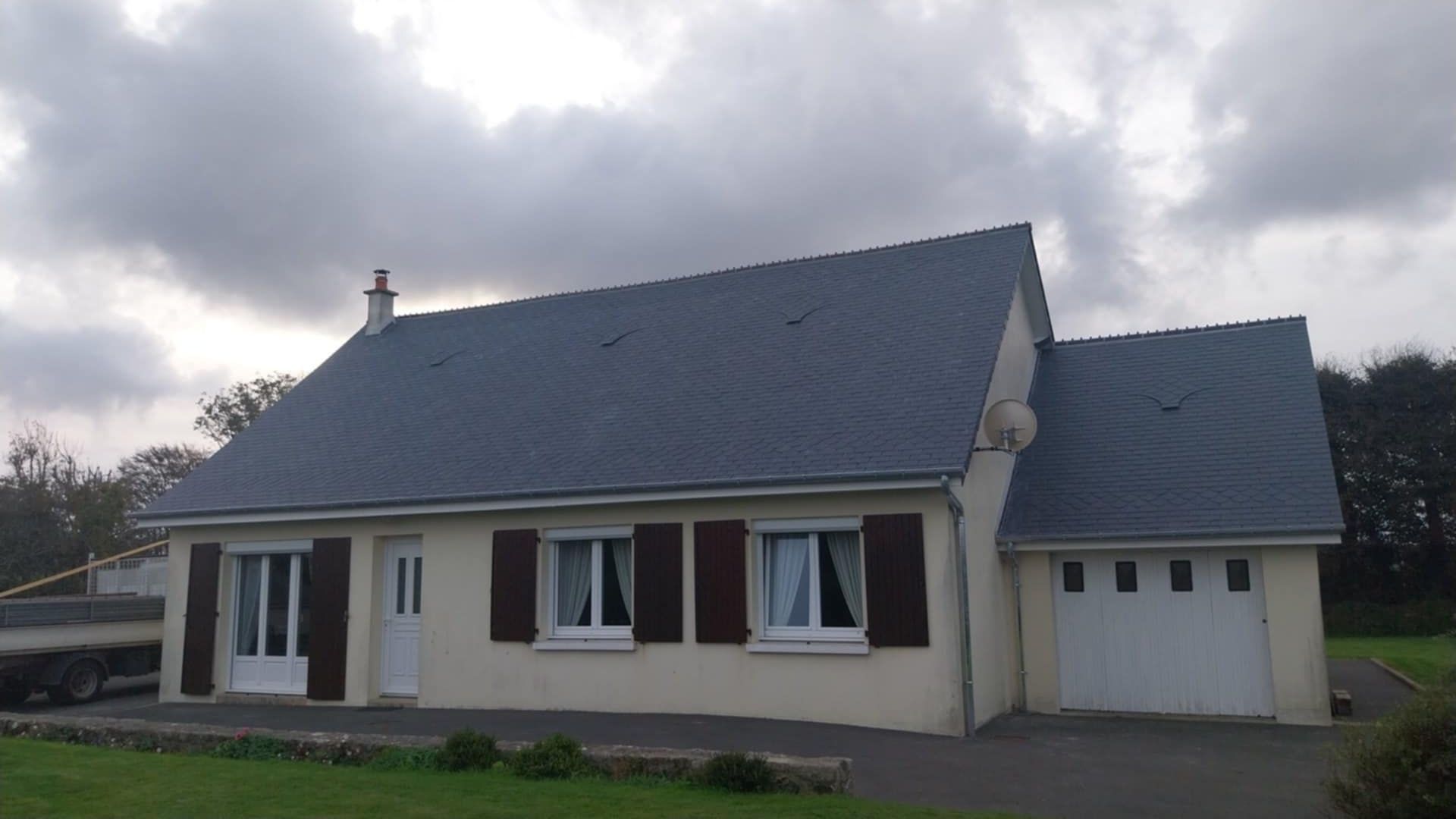 Maison beige avec toit gris, volets marron et garage attenant, sous un ciel nuageux.