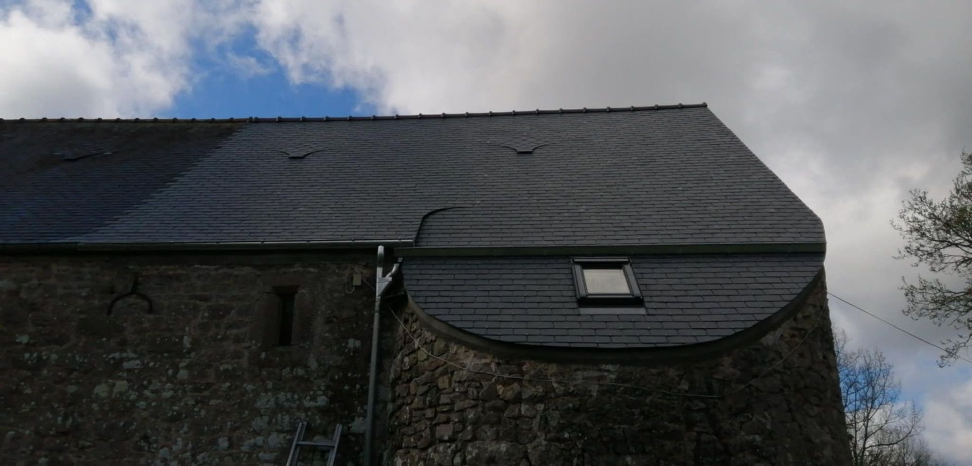 Bâtiment en pierre au toit d'ardoise sombre se détachant sur un ciel nuageux.