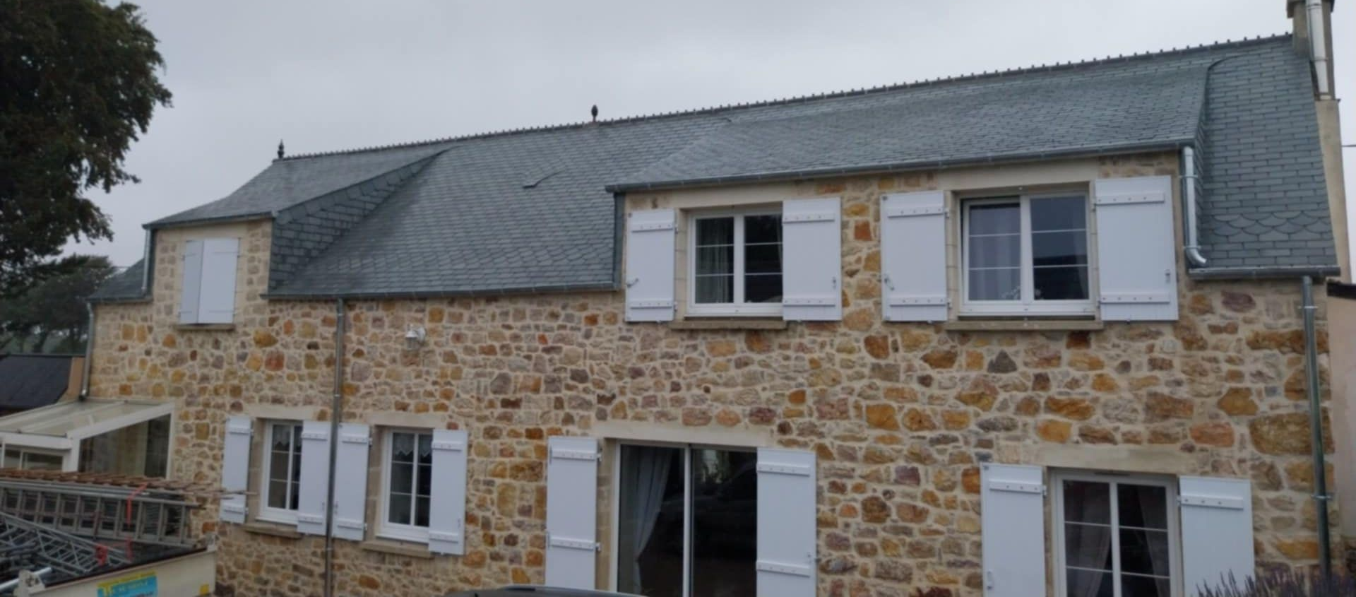 Maison en pierre aux volets blancs, toit gris, ciel couvert.