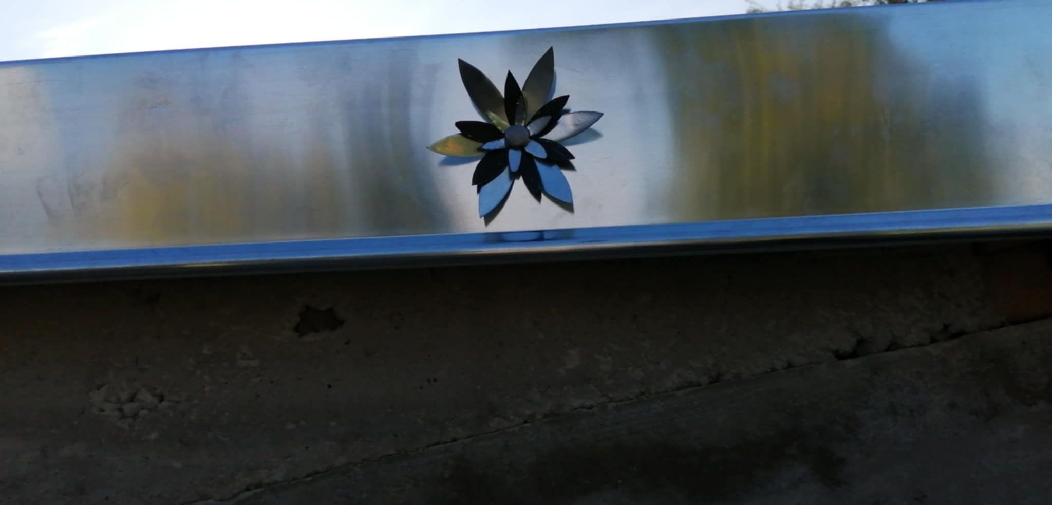 Une fleur décorative en métal sur une surface métallique brillante, reposant sur un socle en béton foncé.