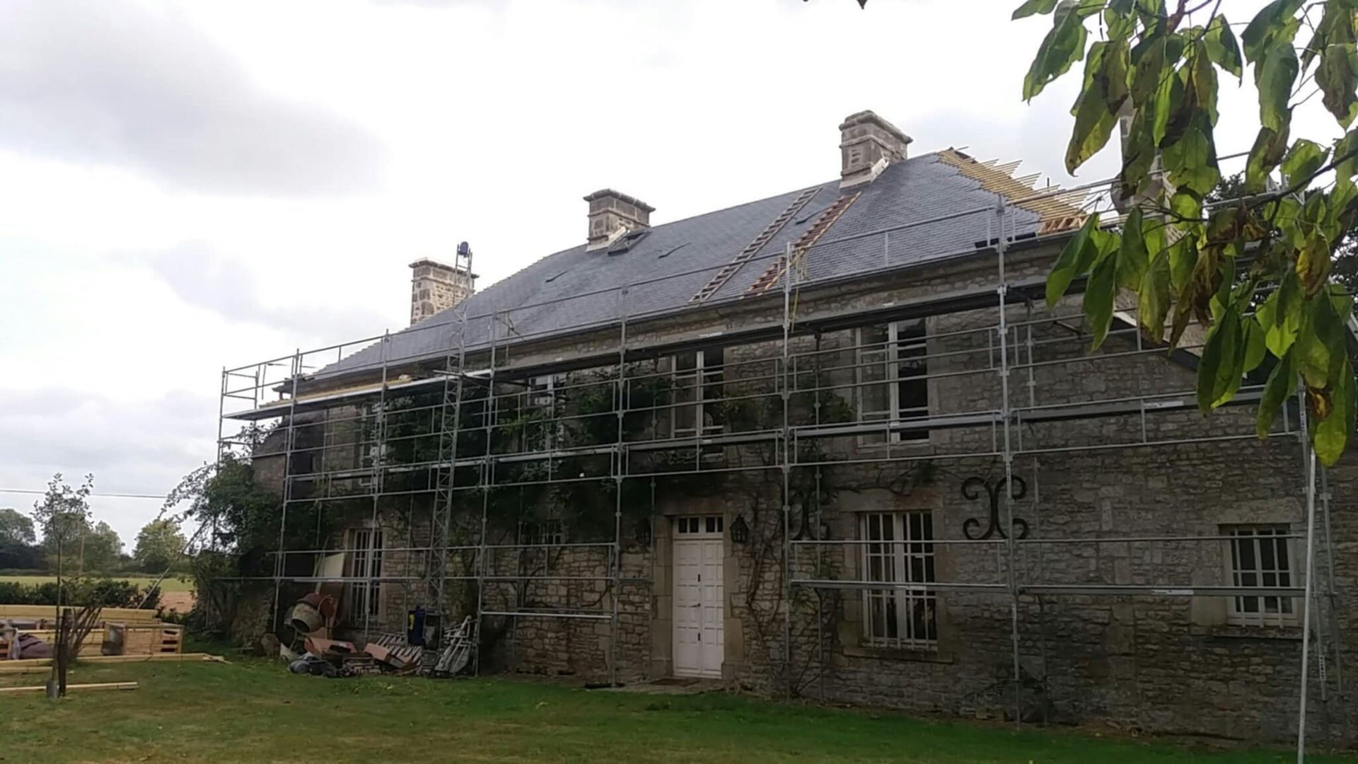 Maison en pierre avec échafaudages, travaux de réparation de la toiture en cours. Ciel nuageux, pelouse verte.