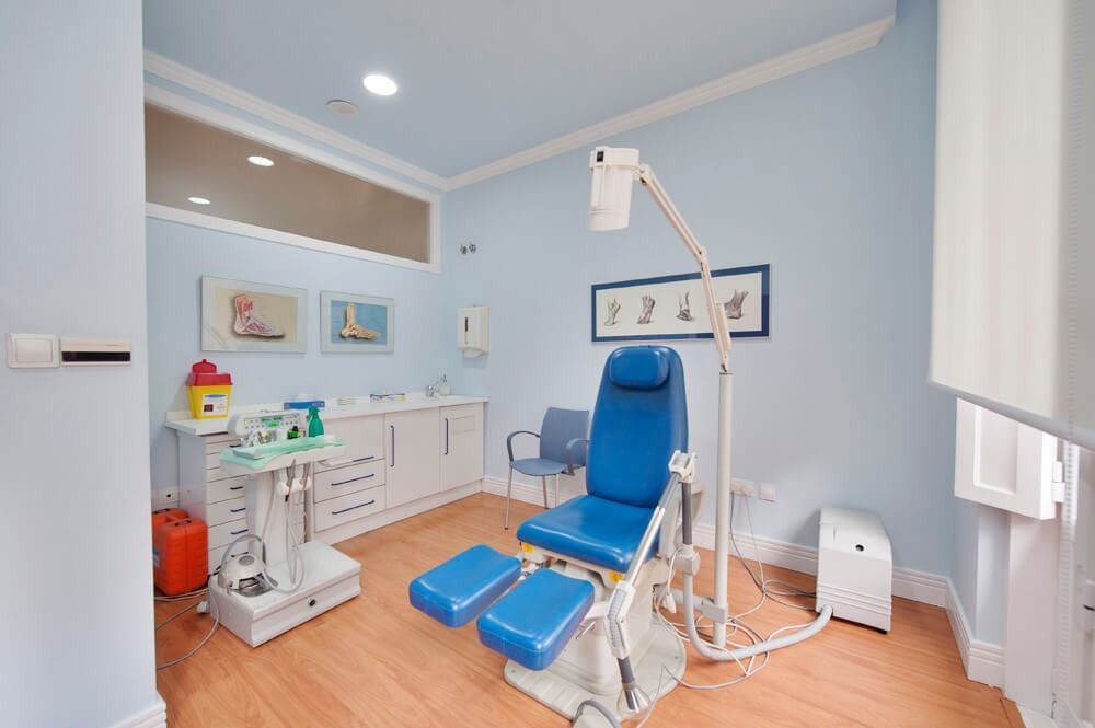 Consultorio de podólogo con silla médica y equipo de color azul, paredes de color azul claro, piso de madera y dos diagramas de pies enmarcados.
