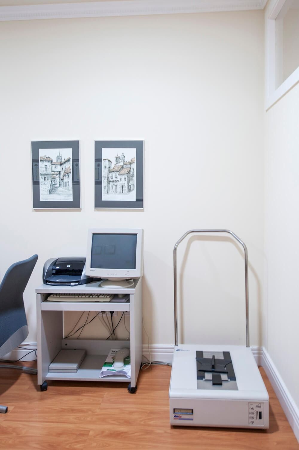 Un rincón de consultorio médico con una computadora, una impresora, una báscula y arte enmarcado en una pared beige.