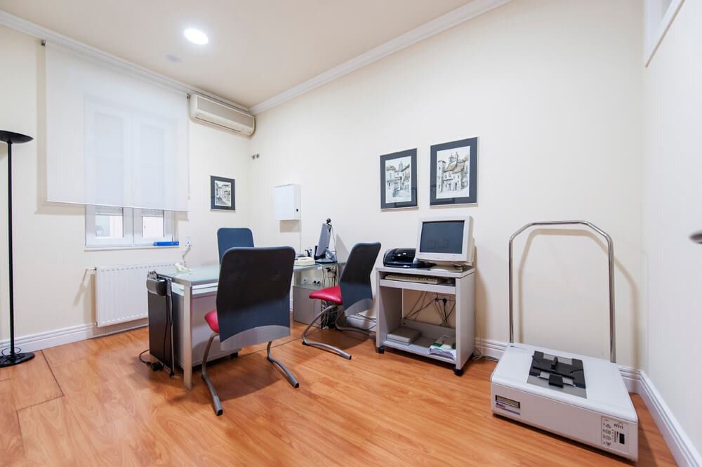 Sala de examen con escritorio, sillas, computadora, equipo médico y obras de arte en la pared.