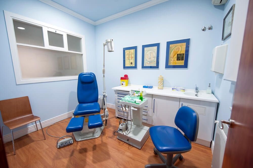 Consultorio médico con paredes azules, silla de exploración, armarios y obras de arte enmarcadas.