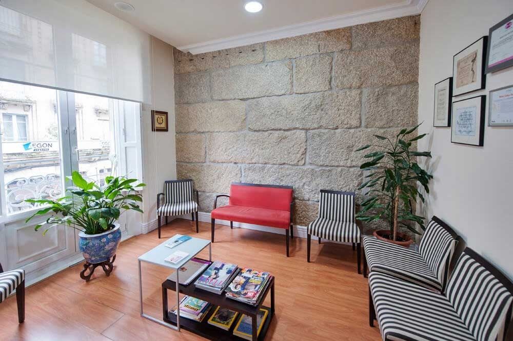 Sala de espera con pared de piedra, asientos rojos y rayados, revistas y una ventana con plantas.