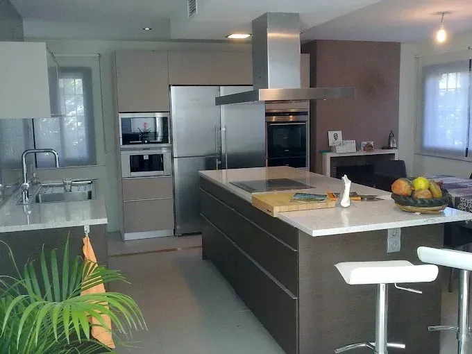 Cocina moderna con isla, taburetes blancos, electrodomésticos de acero inoxidable y plantas en macetas.