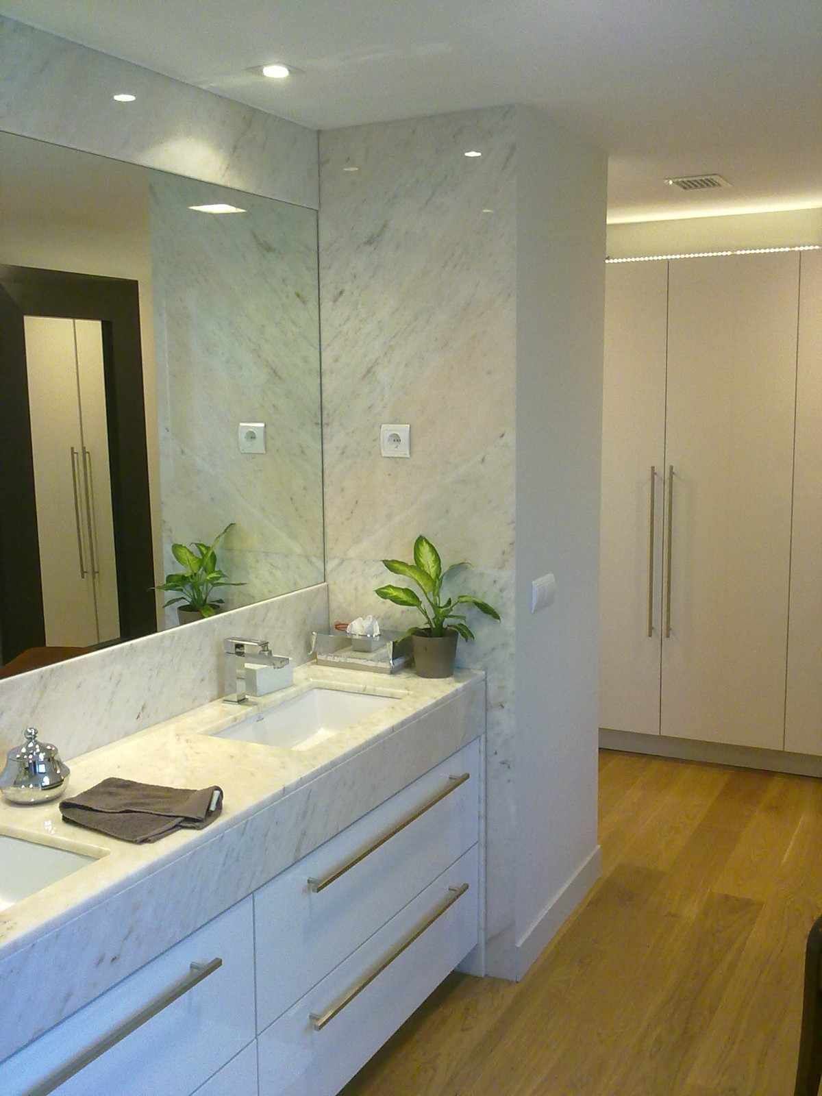Baño moderno con tocador de mármol, espejo grande, lavabo, planta en maceta y armarios empotrados blancos.