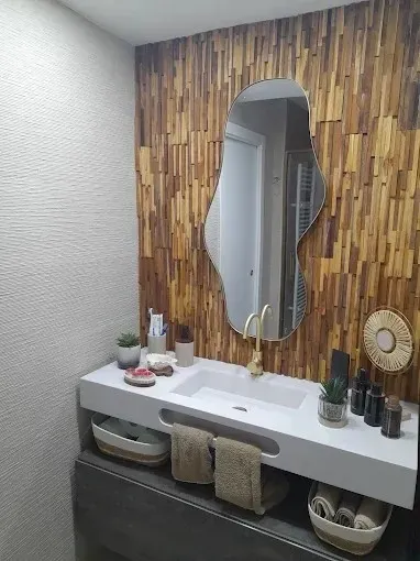 Tocador de baño moderno con pared decorativa de madera, espejo ovalado, lavabo, toallas y elementos decorativos.