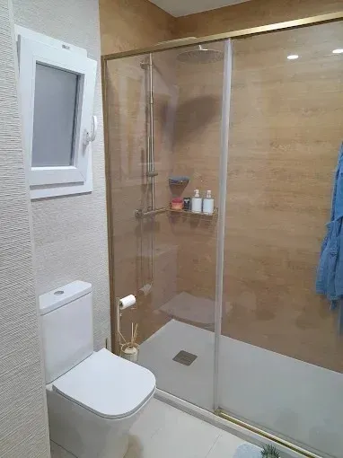 Baño pequeño con inodoro, ducha de cristal, paredes de color beige y una toalla azul colgada en la puerta.