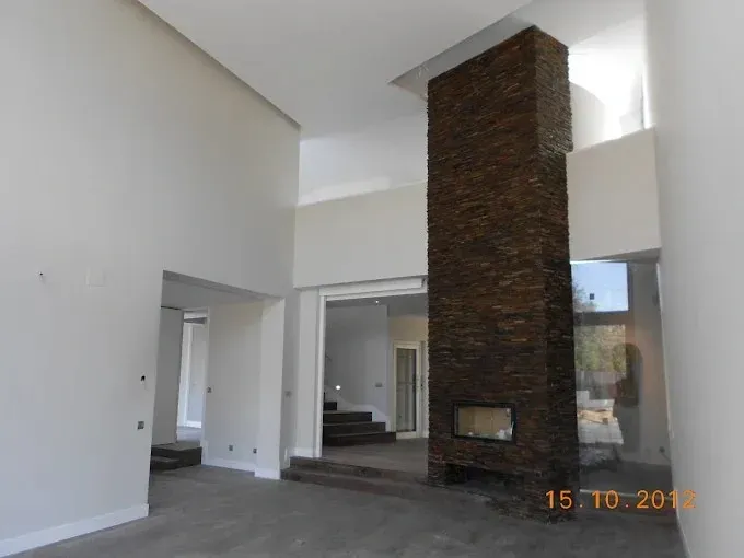 Sala de estar moderna y vacía con una alta chimenea de piedra, paredes blancas y grandes ventanales.
