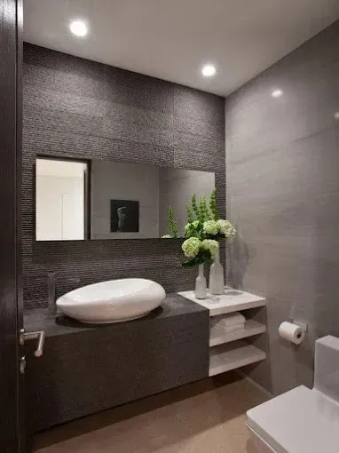 Baño moderno de color gris con tocador flotante, lavabo ovalado, espejo, estantes, flores e inodoro.