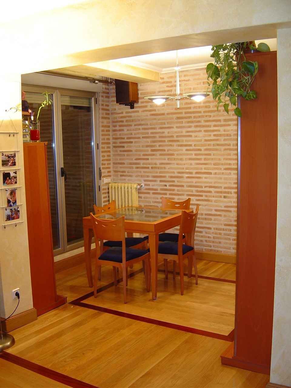 Pequeño rincón comedor con mesa y sillas de madera, pared de ladrillo y planta colgante en una habitación de tonos cálidos.